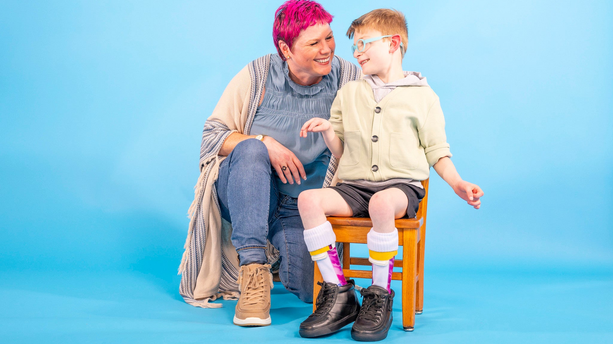 A woman with bright pink hair and a young boy wearing glasses sit together on a small wooden stool against a solid blue studio background. The boy is wearing a light green cardigan, dark shorts, white socks with yellow and pink stripes, and black shoes.