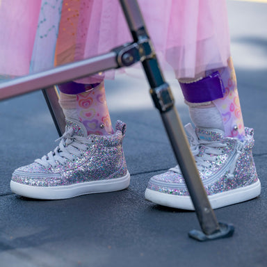 A close-up of a child's feet wearing sparkly silver high-top sneakers with white soles, pink patterned socks, and a pink tutu skirt, standing near a metal walker outdoors.