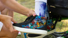 A close-up lifestyle shot of a person holding a black low-top sneaker with a vibrant multicolor floral and skull graphic pattern on the side, while another person sits in a wheelchair nearby.