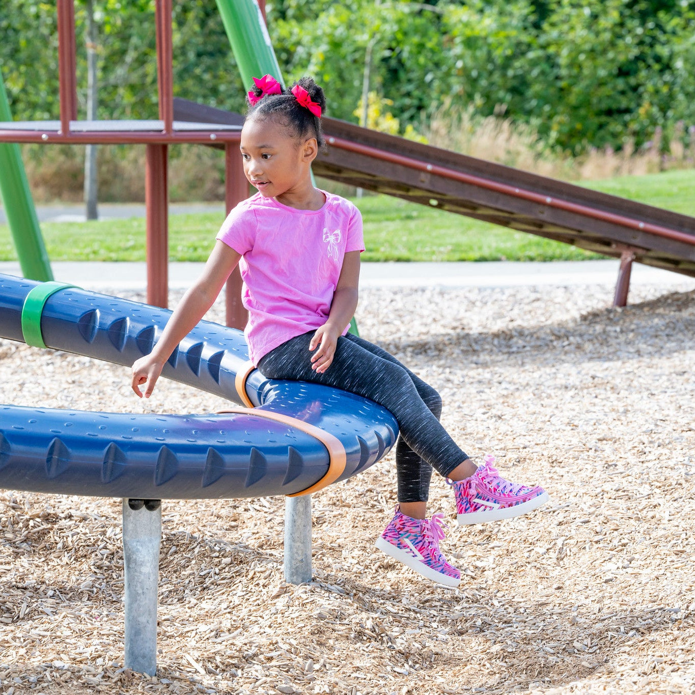 A young girl with pink hair bows sits on a blue curved playground slide at a park. She is wearing a pink t-shirt, dark leggings, and pink sneakers, with a wood chip ground cover and green trees in the background.
