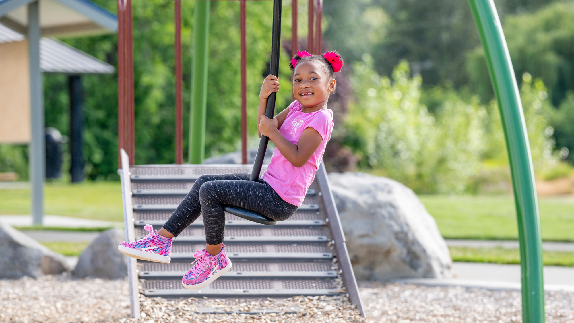 A young girl with pink hair bows sits on a metal playground swing in a park, wearing a pink t-shirt, dark leggings, and pink sneakers.